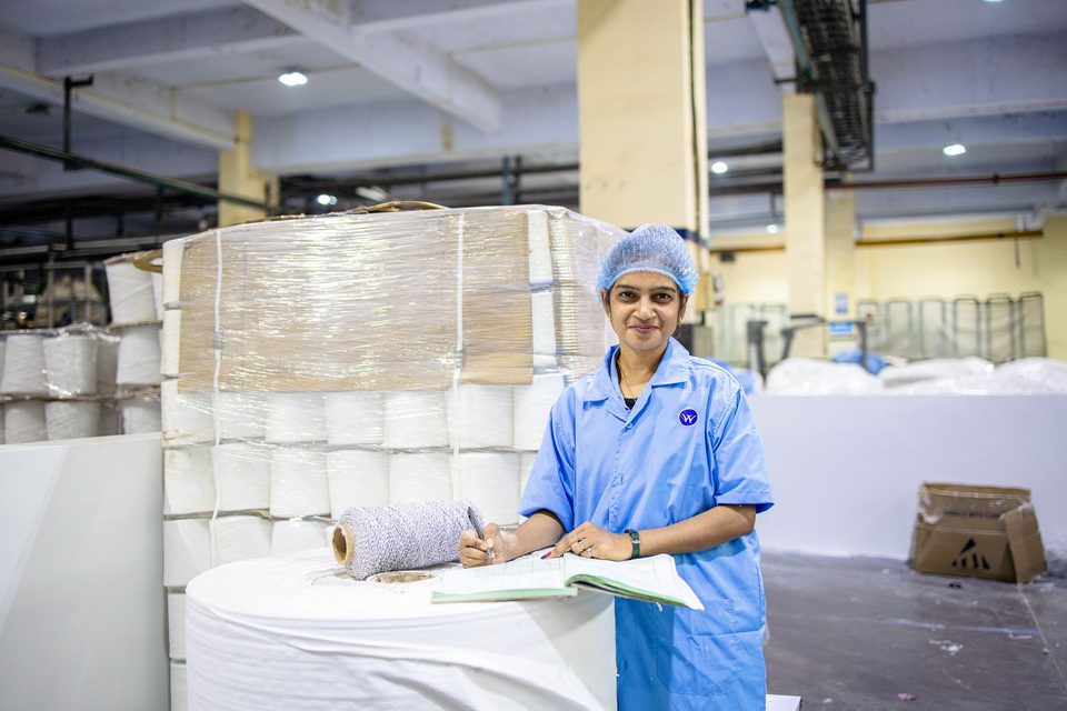A smiling factory worker in a blue coat writing in a register beside spools of yarn